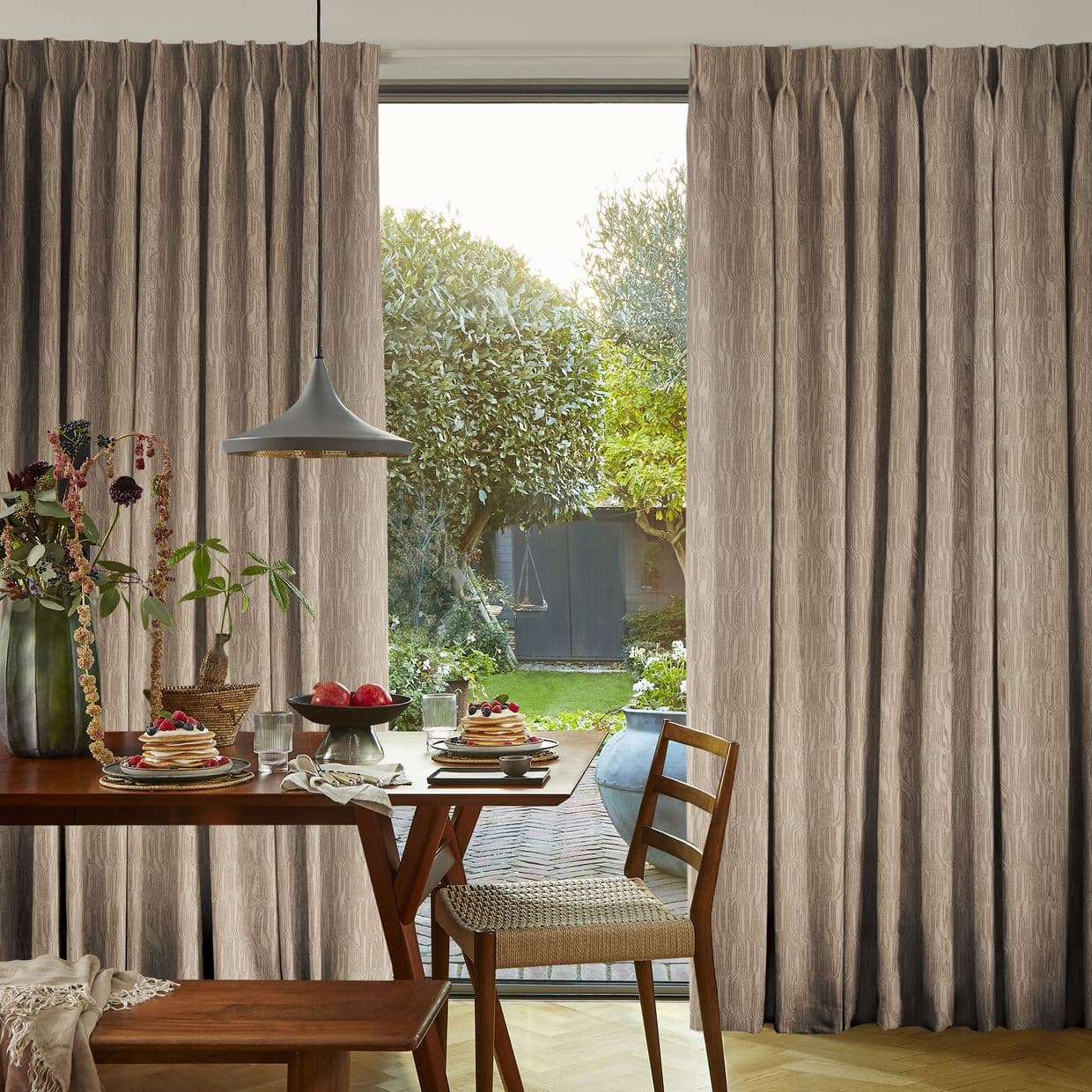 Full-length pleated beige curtains, partially drawn to reveal a center glass door; curtains filter soft daylight into a dining area with wooden table, pendant light and garden view.