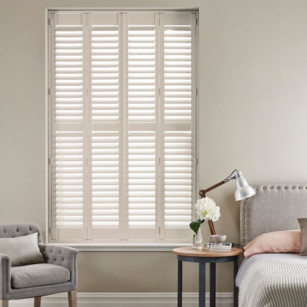 White full-height plantation shutters covering a tall window, louvers tilted to filter soft daylight; set in a neutral-toned bedroom with upholstered bed, bedside table lamp, vase of flowers and armchair.