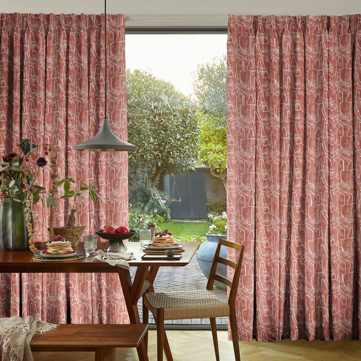 Dusty-rose pleated curtains with abstract white-line pattern, hung full-length and parted; filtering soft morning light and framing a dining table set for breakfast and a leafy backyard garden.