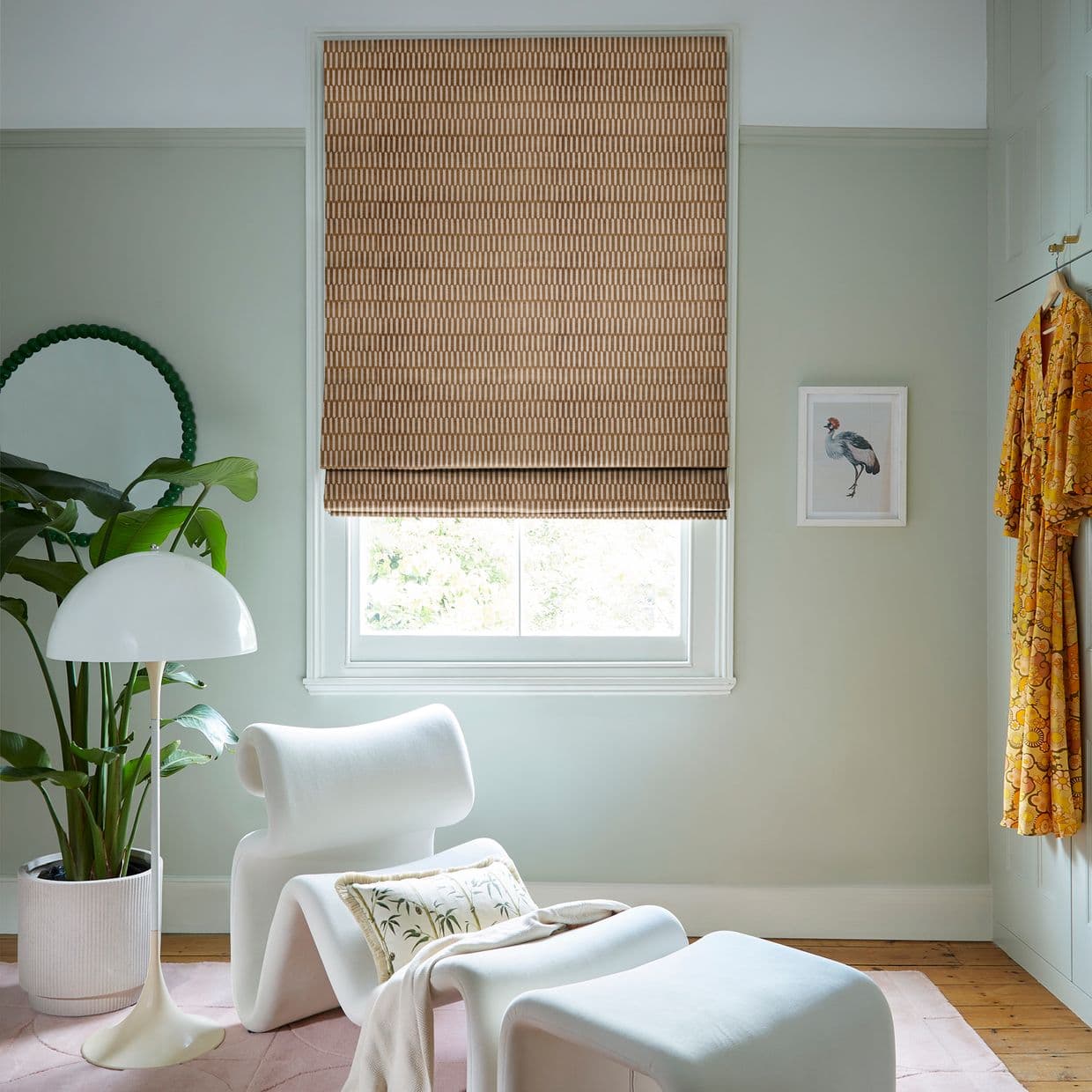 Brown geometric Roman blind, fully lowered across the window, filtering soft daylight; in a pale-green sitting nook with a white lounge chair, floor lamp, potted plant, and framed bird print.