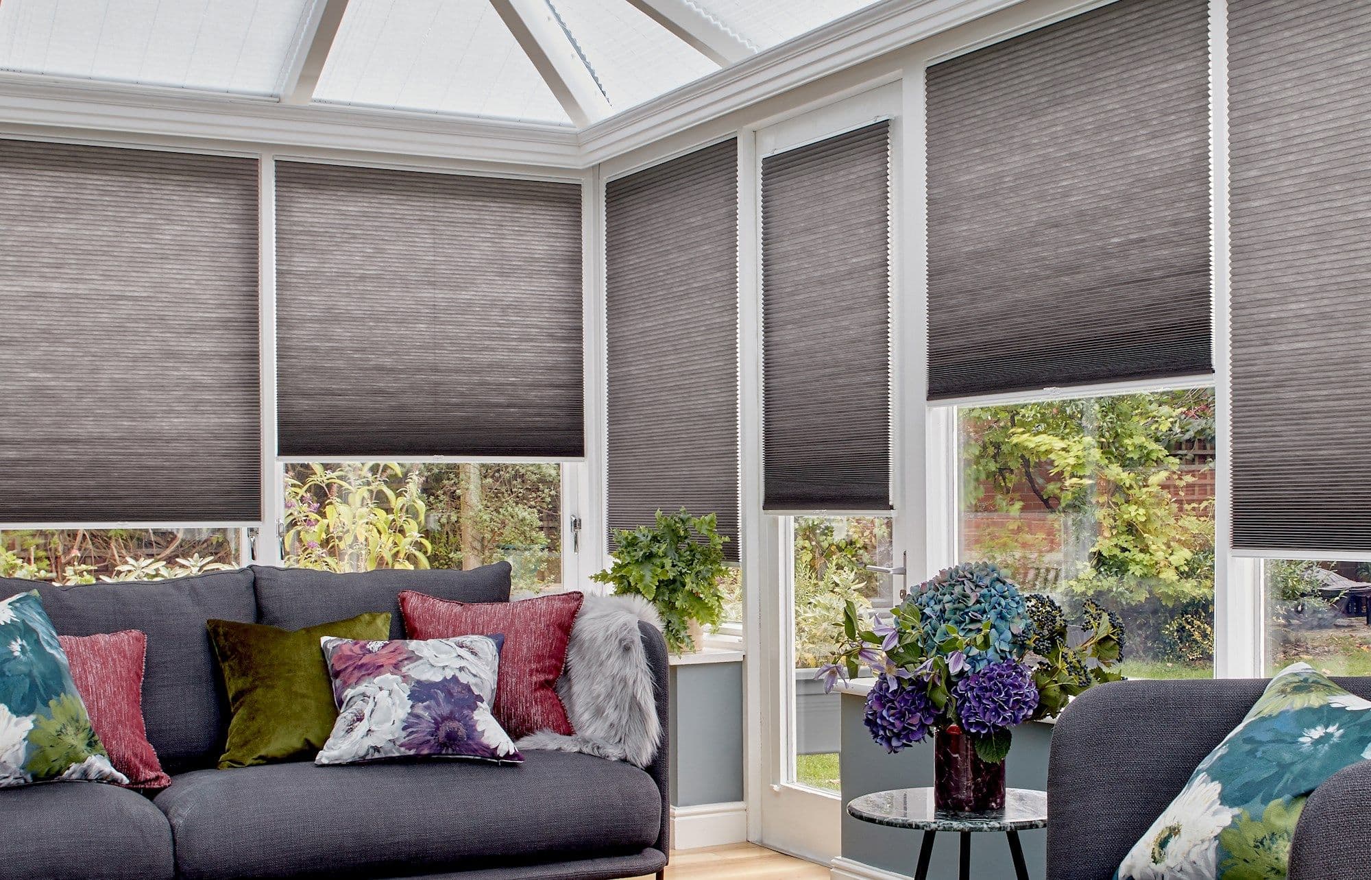 Grey cellular honeycomb blinds, mostly lowered over conservatory windows and glazed door, filtering daylight; bright sunroom with dark grey sofas, colorful floral cushions, potted plants and a garden view.