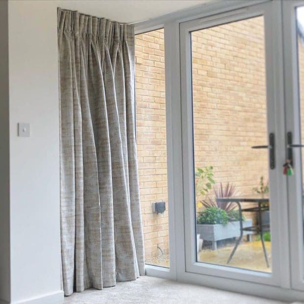 Floor-length pleated grey curtains pulled closed over the left sidelight of glass patio doors, fabric forming folds; bright daytime living room overlooking small brick patio with potted plants and table.
