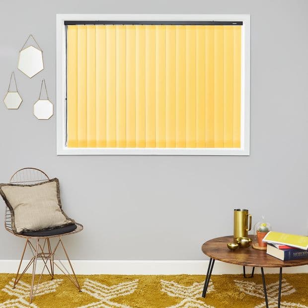Yellow vertical blinds cover a centered rectangular window, closed and softly filtering daylight; set on a grey wall above a mustard rug, wire chair with cushion, round wooden coffee table.