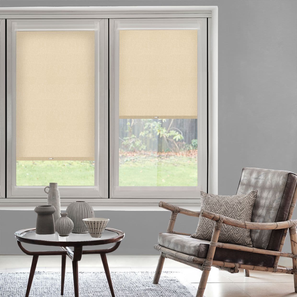 Two beige roller blinds (inside-mounted textured fabric with small pull tabs) â€” left fully lowered, right halfway â€” filtering daylight in a grey-walled living room with rattan armchair and coffee table.