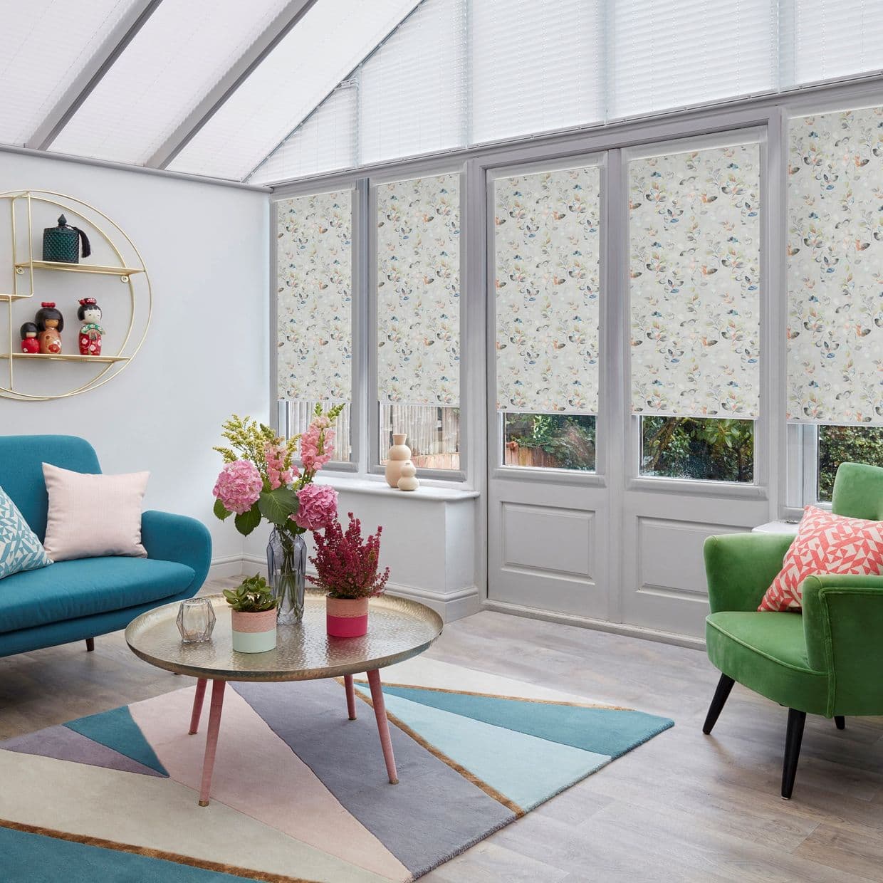 Floral-patterned roller blinds covering tall French windows and doors, lowered to mid-height and filtering light; pleated roof blinds above; bright conservatory with colorful sofas, coffee table and garden view.