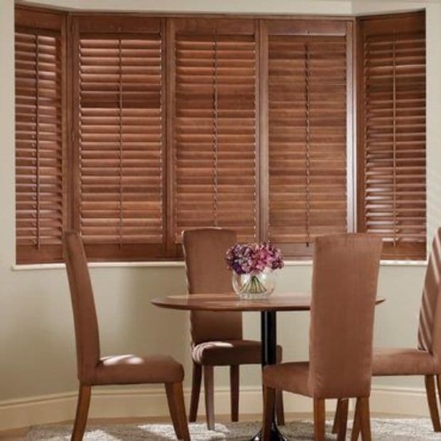 Wooden horizontal blinds (bay window) closed, slats slightly filtering light; a small dining area with round table, four brown upholstered chairs and glass vase of purple flowers against neutral walls.