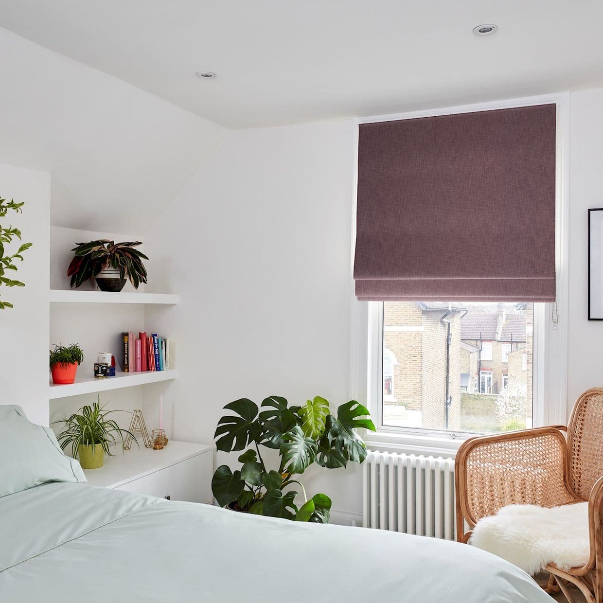 Mauve Roman blind, lowered over a tall window with fold detail, filtering daylight; in bright white bedroom with built-in shelves, plants, rattan chair and radiator, views of nearby brick houses.