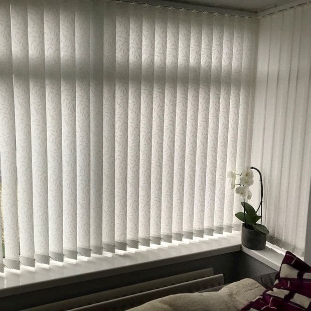 White vertical blinds with subtle leaf pattern, fully closed over an L-shaped bay window, filtering soft daylight; windowsill holds a potted white orchid, radiator below, blanket and cushion nearby.