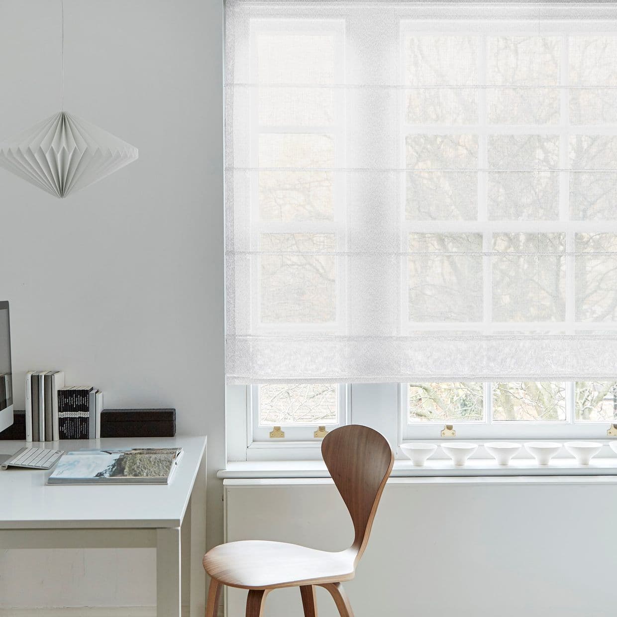 A white sheer Roman shade lowered over a multi-pane window, softly filtering daylight and revealing tree silhouettes; minimalist home office with white desk, wooden chair, pendant lamp and soft natural light.