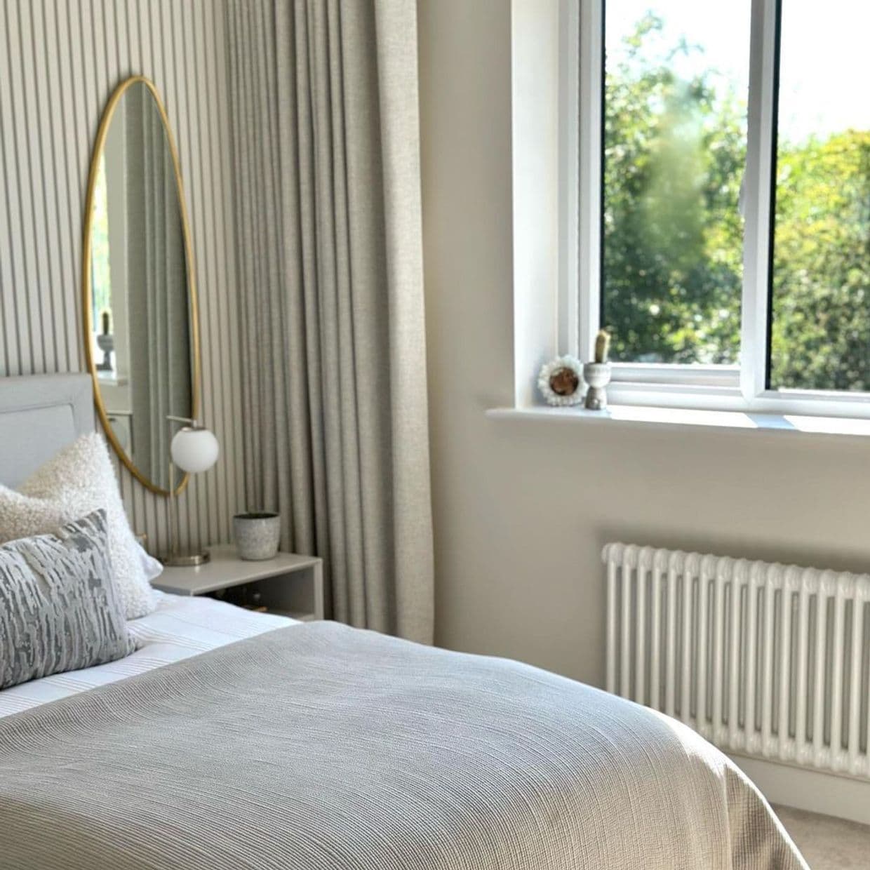 Full-length beige pleated curtains, floor-to-ceiling and drawn to the left, drape in soft folds, gently filtering daylight from an adjacent open window in a neutral, sunlit bedroom with garden view.