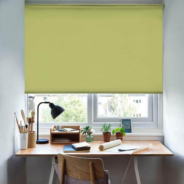 A pale-yellow roller blind, lowered across the upper window with visible metal headrail, filtering daylight. It shades a small home workspace: wooden desk, lamp, potted plants and a tree-lined view.