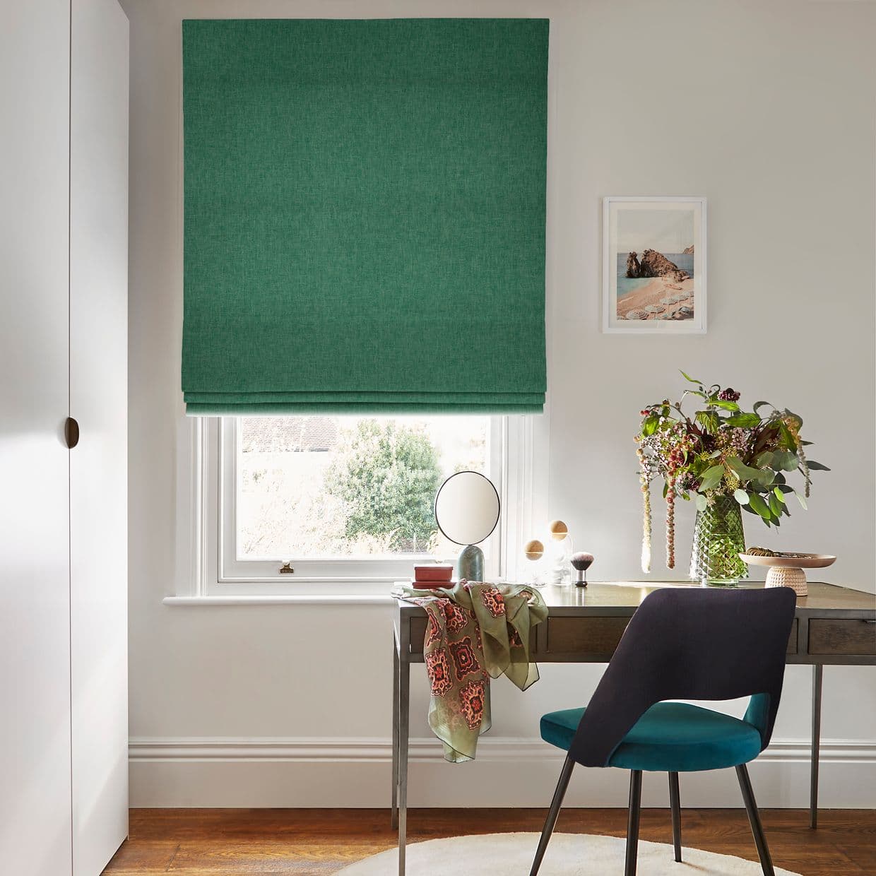 Green fabric Roman blind, lowered with soft horizontal folds, filtering daylight through a white-framed sash window; sits above a wooden desk with chair, mirror, vase of flowers and neutral walls.