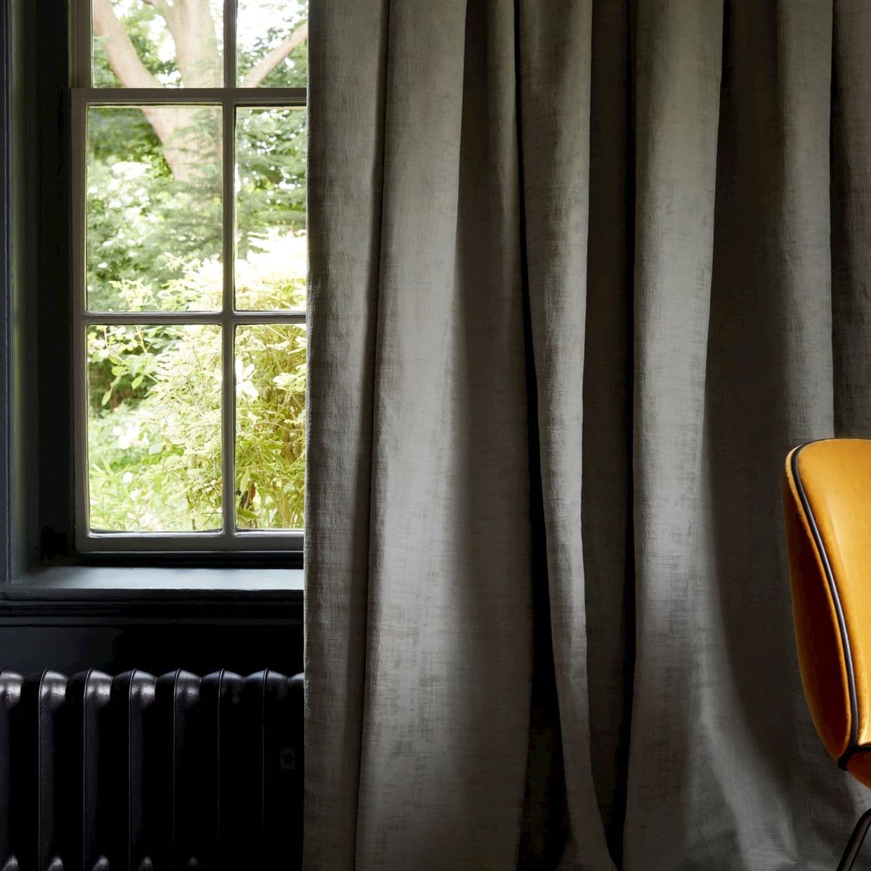 Heavy grey linen curtains, floor-length, pleated, drawn mostly closed across a sash window with one side open, filtering daylight into a dim room with radiator and orange chair; garden outside.