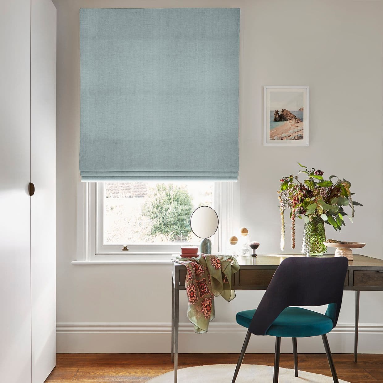 Light-blue fabric Roman blind, lowered with gentle folds; filtering soft daylight through a small sash window; above a desk with chair, mirror and vase of flowers in a home office.