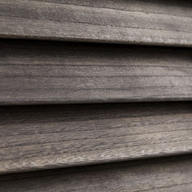 Horizontal wooden blinds, narrow slatted wood shown in close-up; closed and overlapping, textured grain visible; filtering/diffusing light in a dim interior setting with neutral grayâ€‘brown tones.