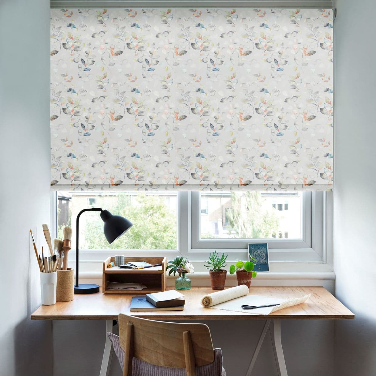 Object: lowered floral roller blind with watercolor leaves covering the window; Action: filtering diffuse daylight; Context: tidy home desk with lamp, art supplies, succulents and a tree-lined suburban view.