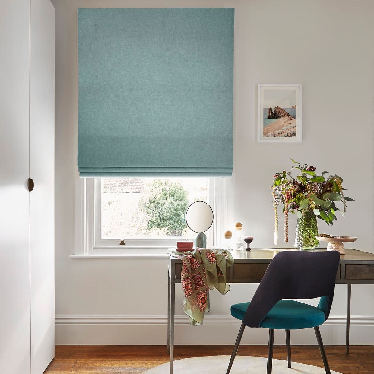 A teal fabric Roman blind, lowered with neat horizontal folds, filtering daylight over a white-framed window; a desk with mirror, vase of flowers and teal chair in a bright study.