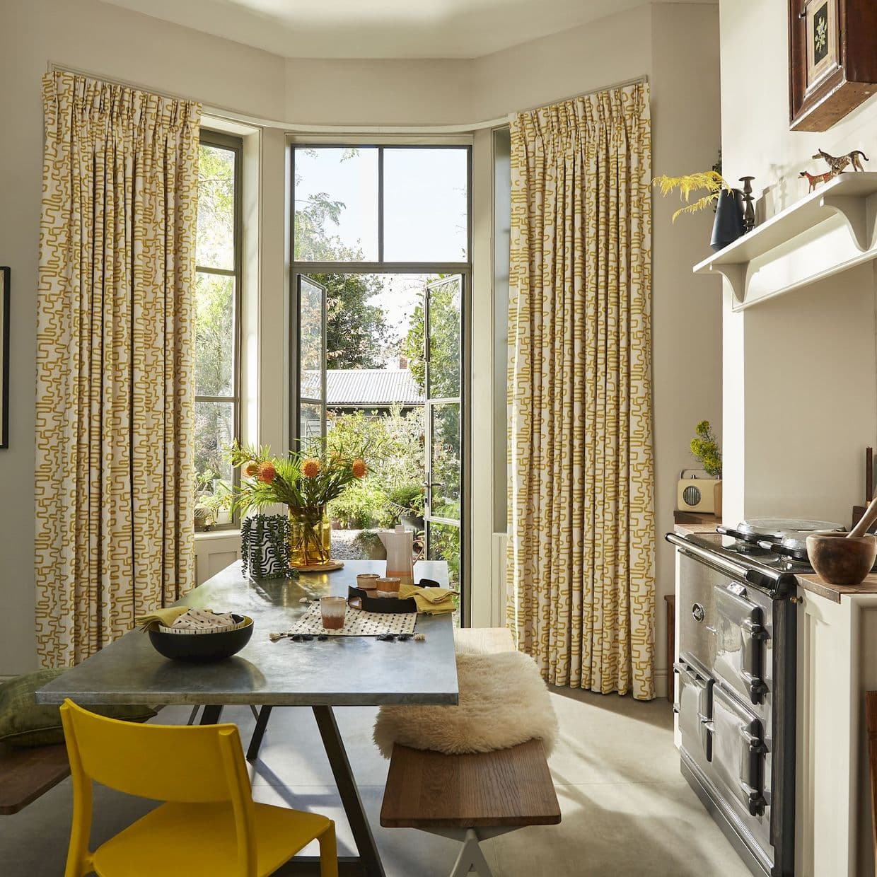 Full-length curtains, yellow geometric pattern, hung at bay window sides; pulled back to frame and admit sunlight through open doors into a bright kitchen with table, bench and garden view.