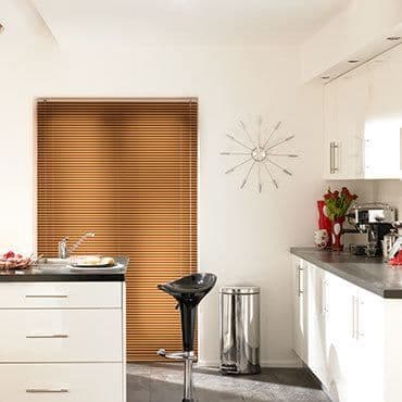 Brown wooden horizontal blinds lowered over the window, closed and filtering daylight into a modern white kitchen with black counters, sink, bar stool, stainless bin, wall clock and red tulips.