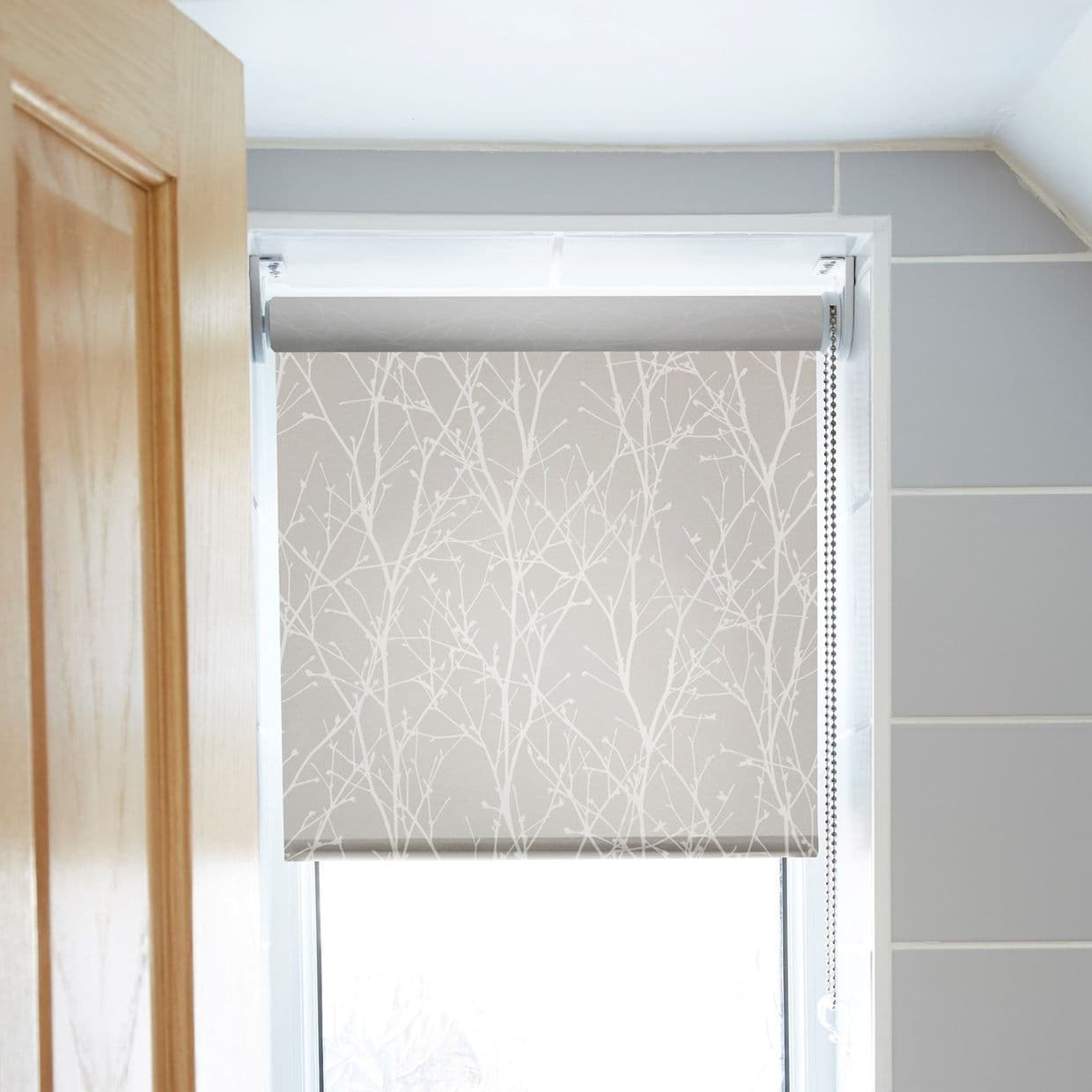 A grey roller blind with white twig pattern and right-side bead chain is partially lowered, filtering daylight through a window frame in a tiled, neutral-toned room beside a wooden door.