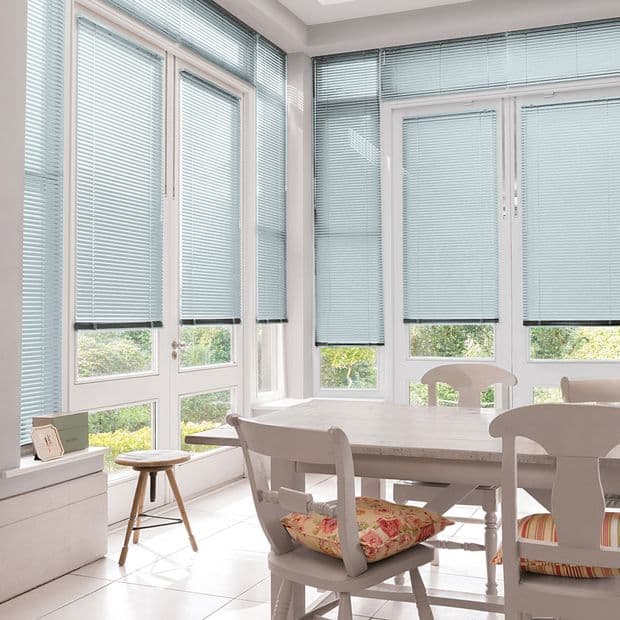 Light-blue horizontal Venetian blinds cover tall French doors and surrounding windows, lowered and filtering soft daylight; bright white dining nook with wooden table, chairs, floral cushions, tiled floor, garden beyond.