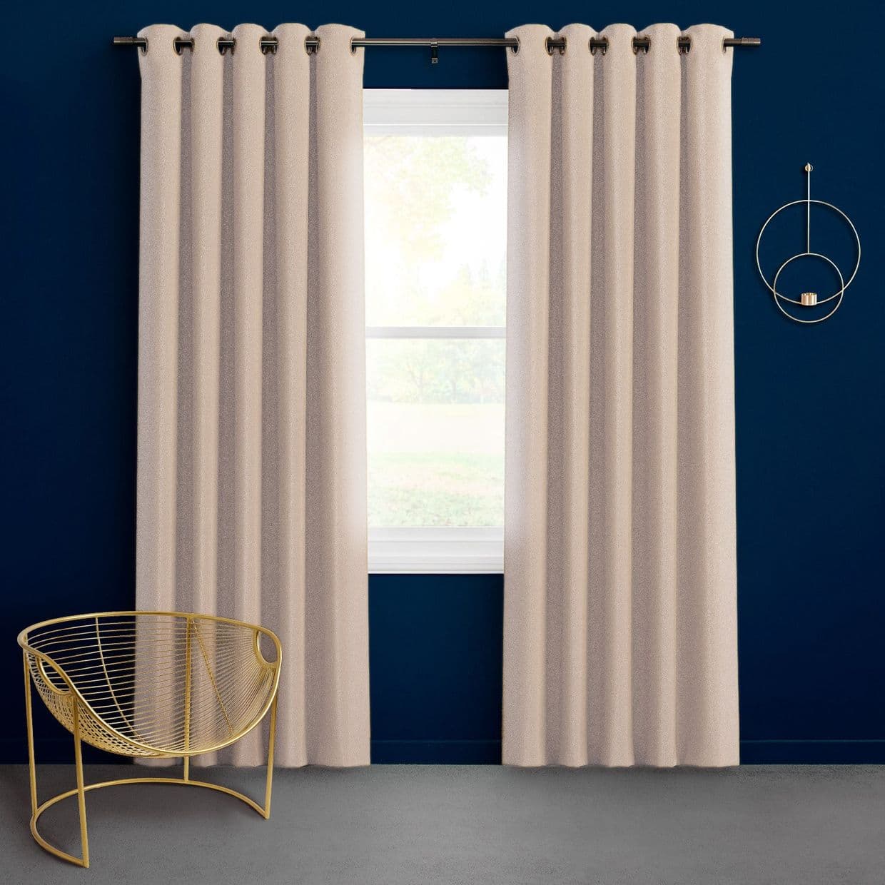 Textured beige grommet-top curtains on a metal rod, slightly parted to reveal a bright window filtering daylight; against a deep navy wall with a gold wire chair.
