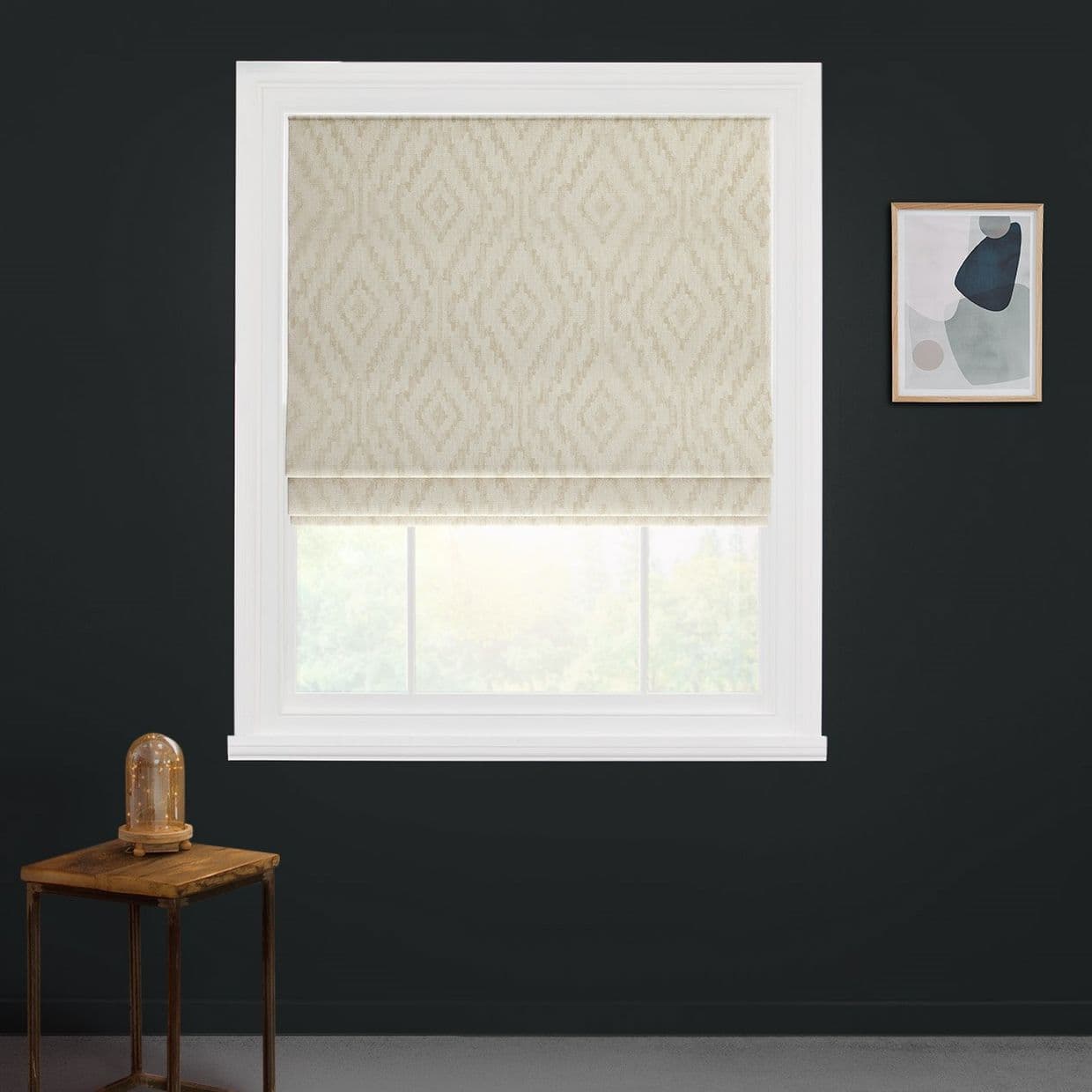 Beige patterned Roman shade lowered, filtering soft daylight through white-framed window set in a dark charcoal wall, with a small wooden side table and a framed abstract print nearby.