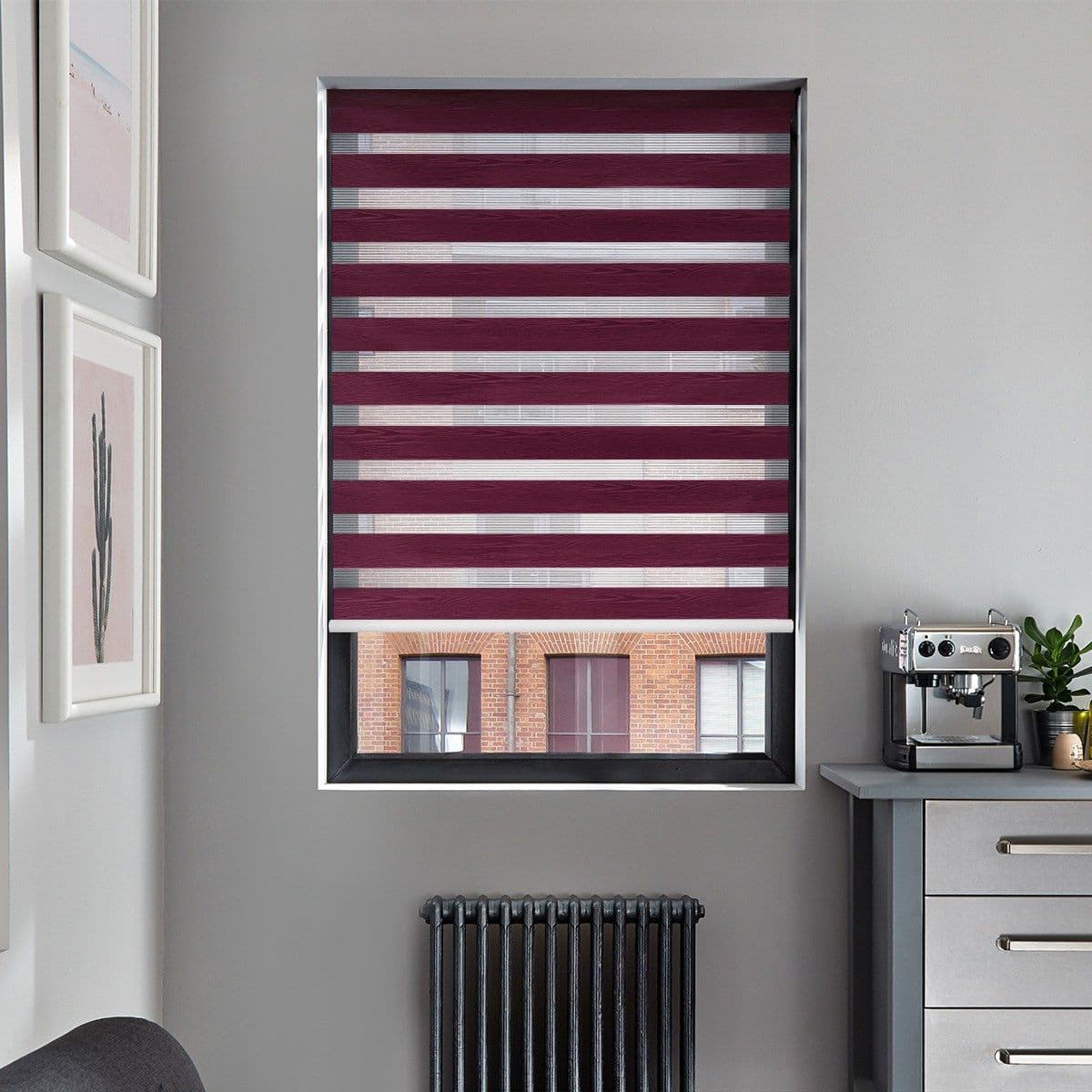 Dual-layer zebra roller blind (burgundy opaque and sheer horizontal stripes) fully lowered across window, filtering light and revealing partial view of a brick building; gray kitchen corner with radiator.