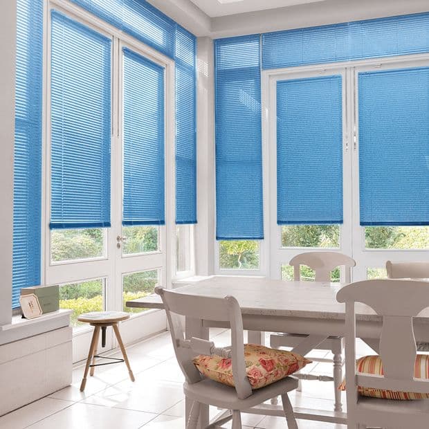 Blue pleated window blinds, fully lowered across tall corner windows, filtering bright daylight; set in a sunlit white dining area with wooden table, chairs, tiled floor and garden view.