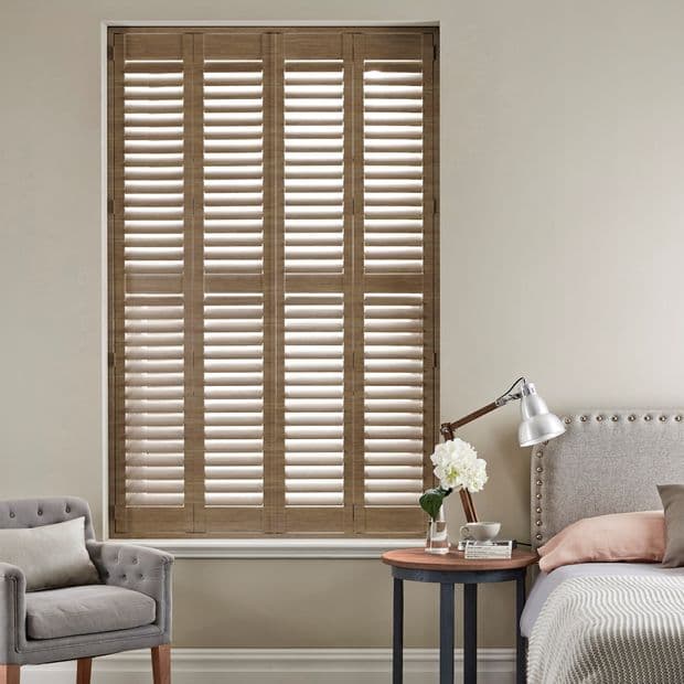 Full-height wooden plantation shutters with four panels and louvers, closed with slats tilted to filter daylight; set in a neutral-toned bedroom beside a tufted armchair, bed, and bedside table lamp.