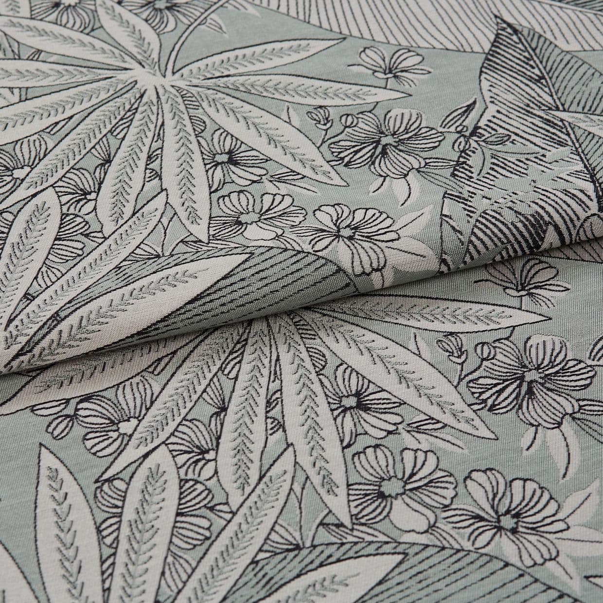 Folded floral-patterned fabric with leaf and flower print positioned diagonally, layered and partially overlapped; black outlined botanical motifs on a pale mint background, photographed close-up in soft lighting.