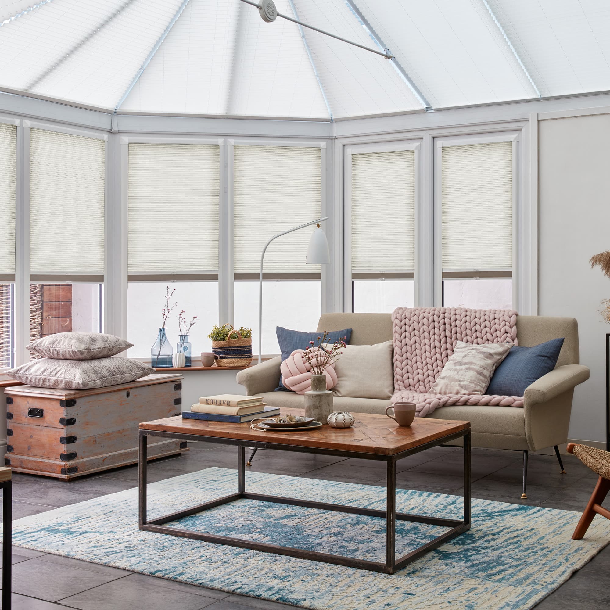 Pleated cellular blinds over bay windows and roof, lowered and softly filtering daylight in a sunlit conservatory with a beige sofa, chunky pink throw, wooden coffee table and blue rug.