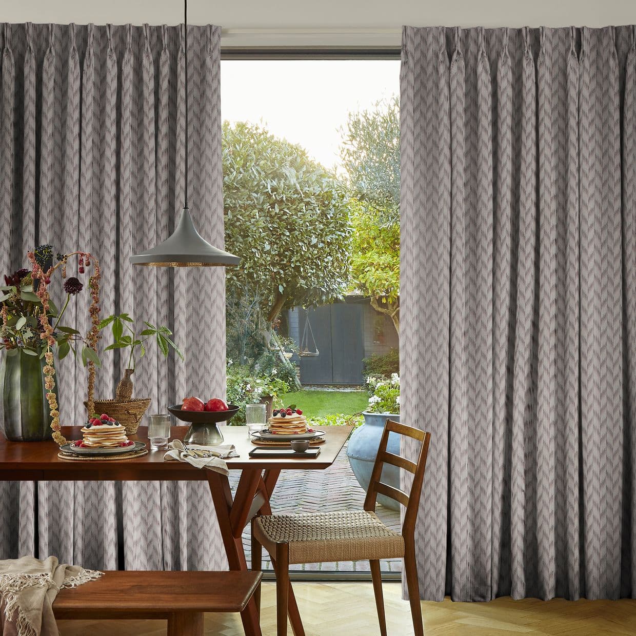 Full-length pleated grey patterned curtains, parted to reveal sliding glass door, filtering daylight; inside, wooden dining table set for breakfast with pendant lamp; outside, a green garden and small shed.