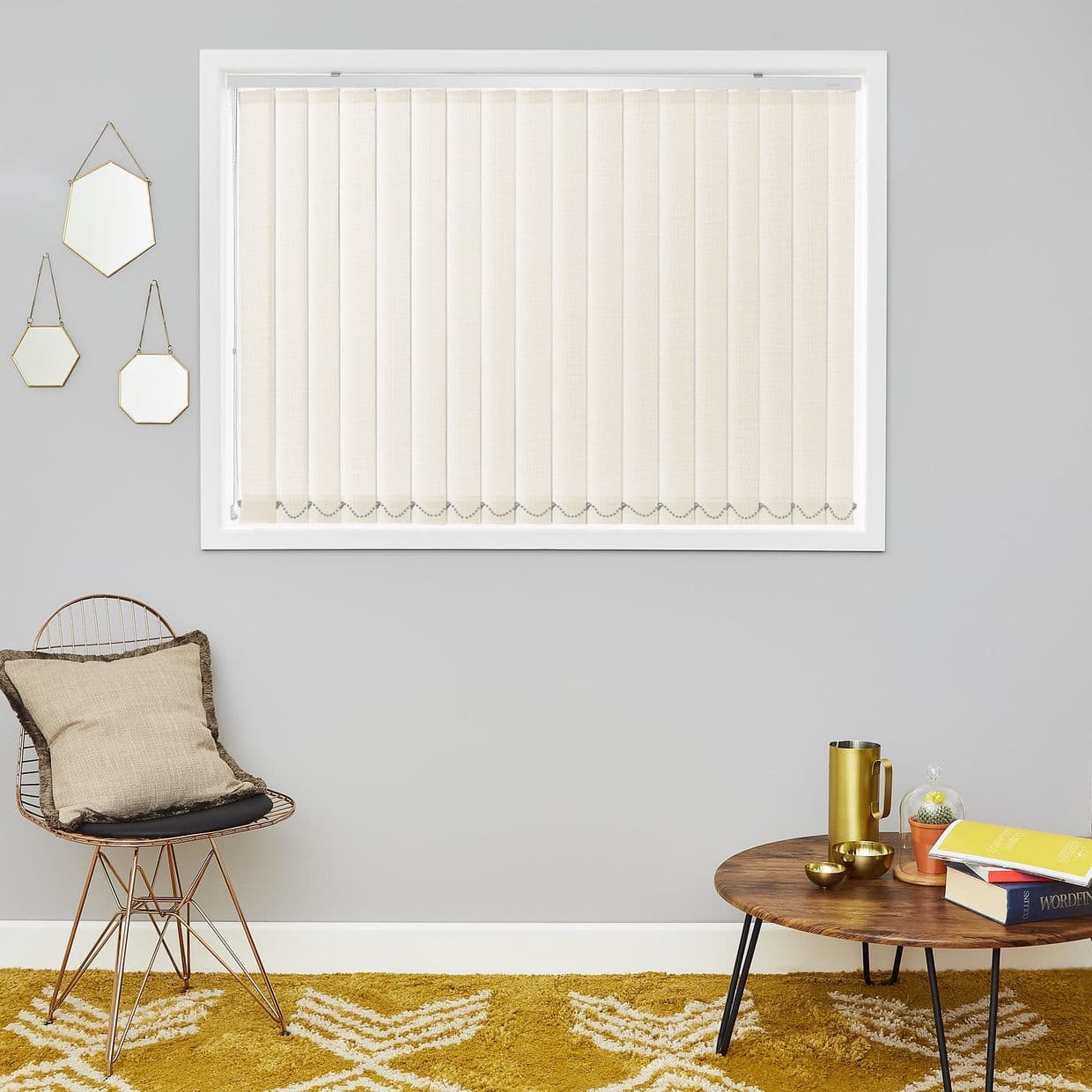 Beige vertical blinds, inside-mounted in a white frame with scalloped bead-chain bottoms, closed and diffusing daylight; set on a grey living room wall above a wire chair, coffee table, mustard rug.