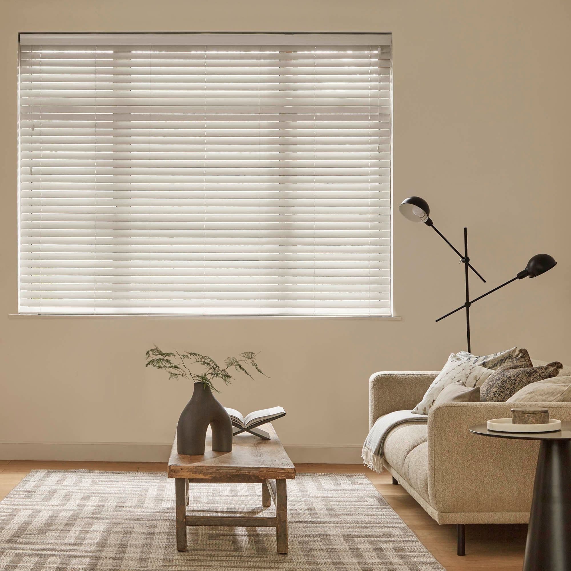 White horizontal blinds covering a large rectangular window, fully lowered with slats closed, softly filtering daylight; in a neutral living room with beige walls, sofa, wooden coffee table and lamp.
