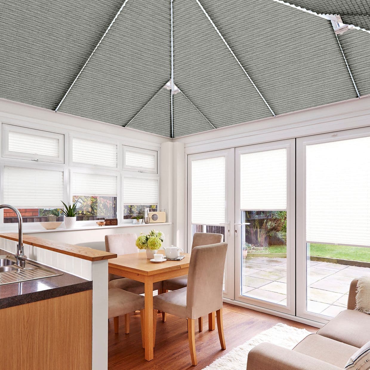 Pleated grey conservatory roof blinds, radiating from the apex, are fully closed, diffusing daylight; matching white pleated window and door shades in a kitchen-dining conservatory overlooking a patio and lawn.