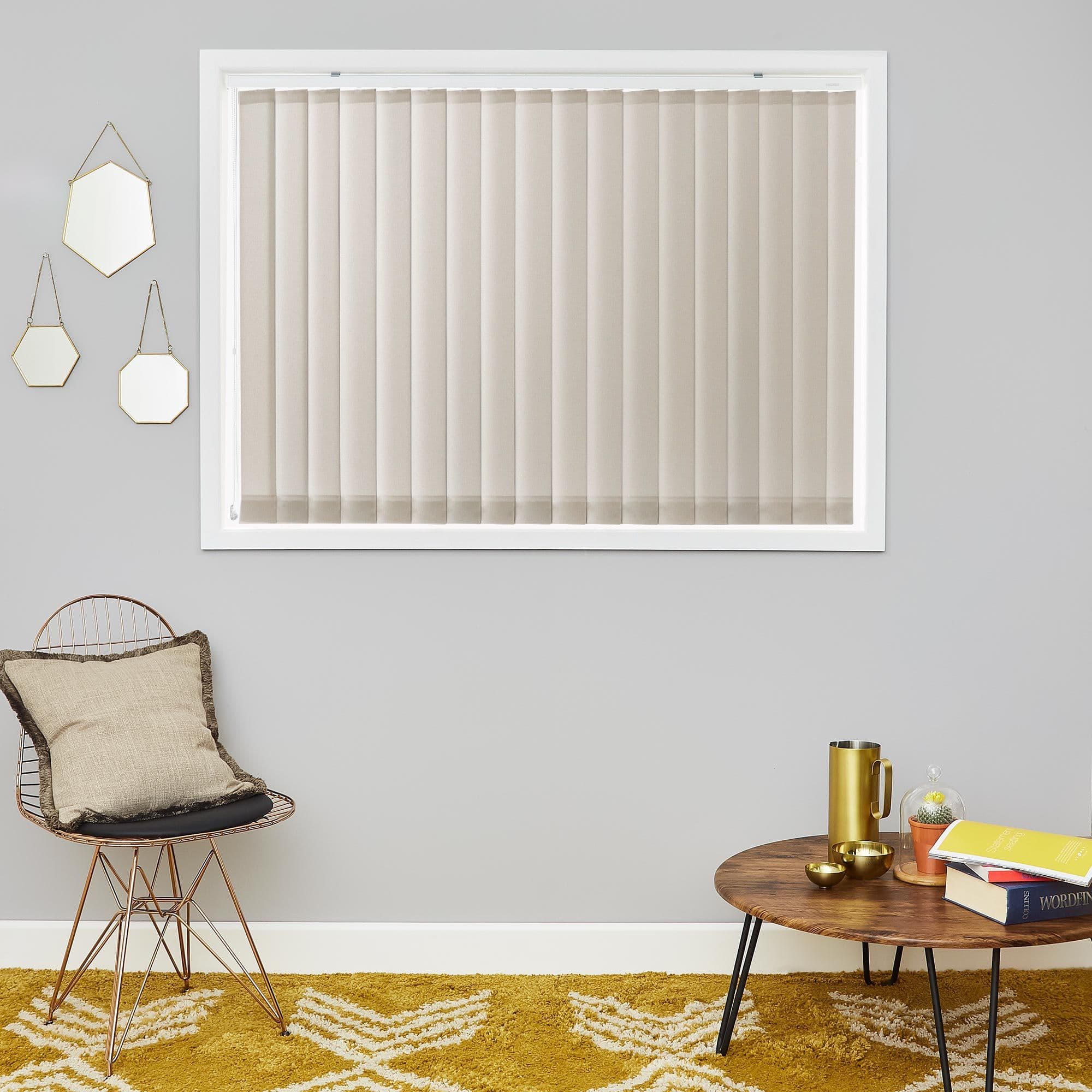 Beige vertical blinds fully closed across a rectangular window, filtering soft light; framed by pale gray wall with hexagonal mirrors, wire chair with cushion, wooden coffee table and mustard rug.