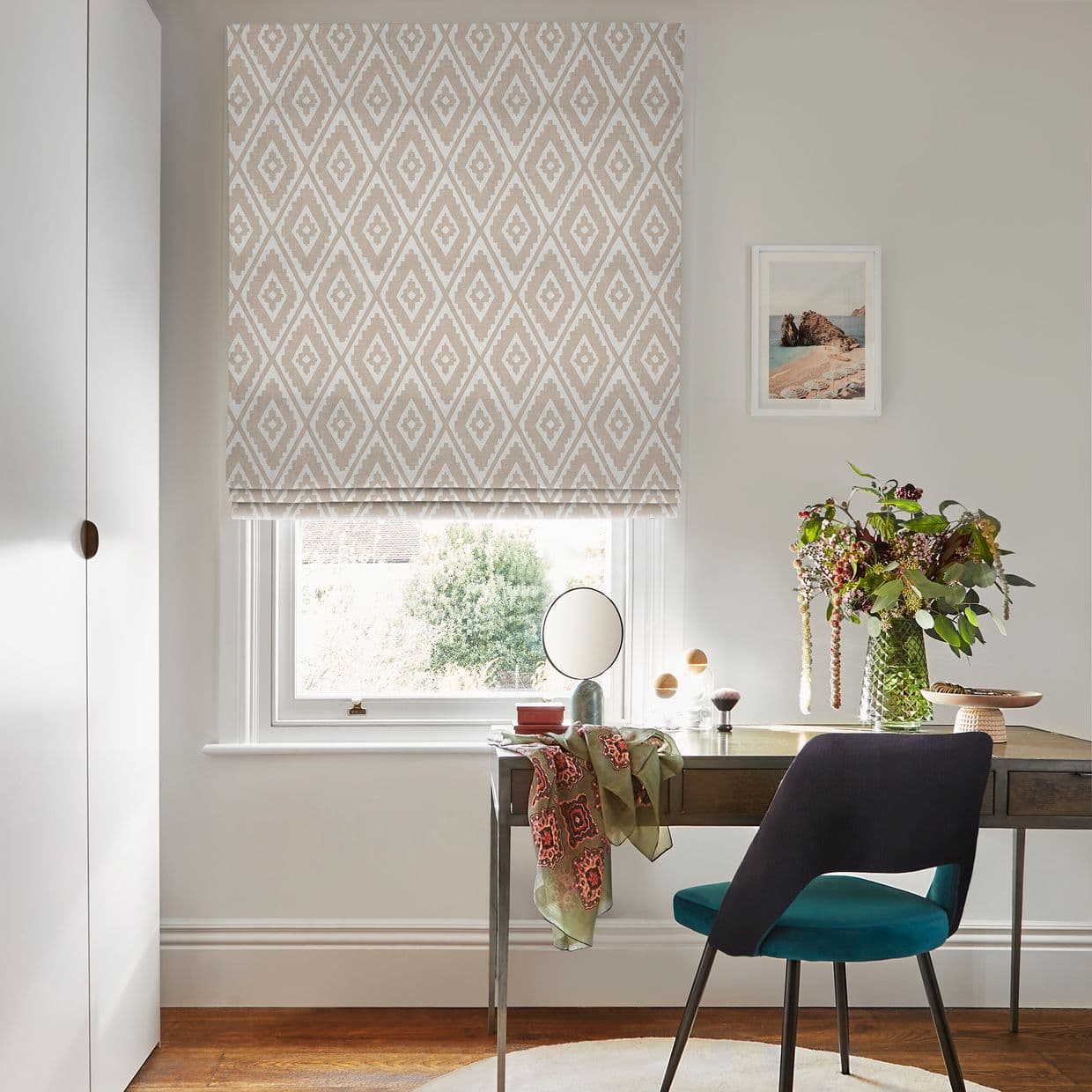 Beige-and-white diamond-pattern Roman blind lowered over the window, softly filtering daylight; a desk below holds a vase of flowers and mirror, teal chair, pale walls and framed print.
