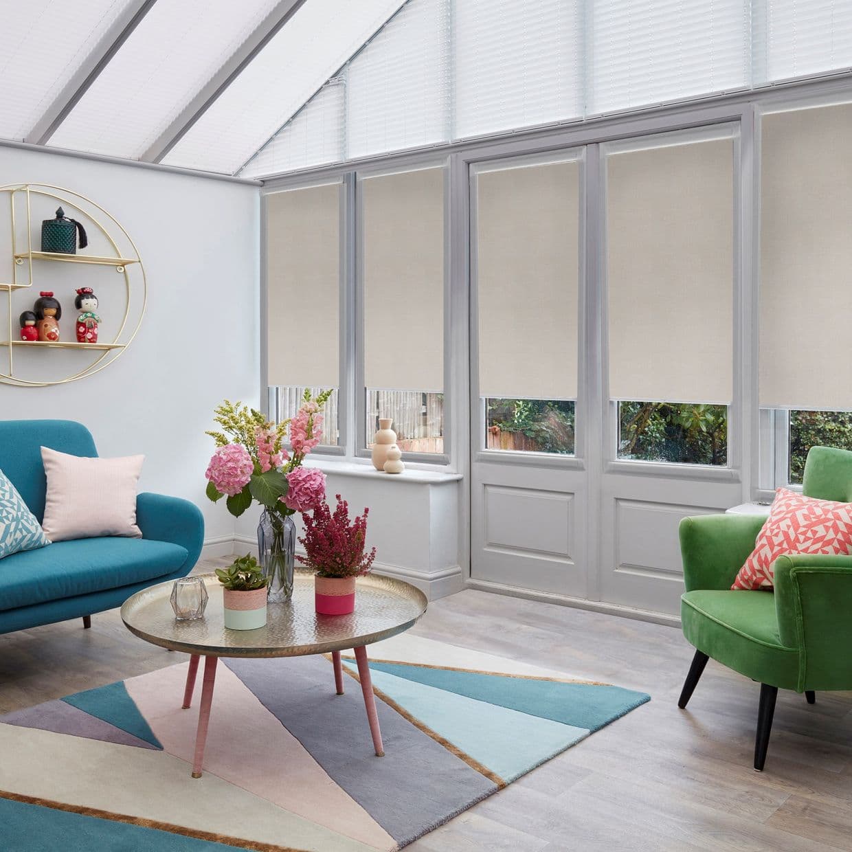 Beige roller blinds covering tall glass doors and windows, lowered halfway and filtering daylight; conservatory roof has pleated blindsâ€”bright sunroom with colorful sofas, a coffee table, and potted flowers.