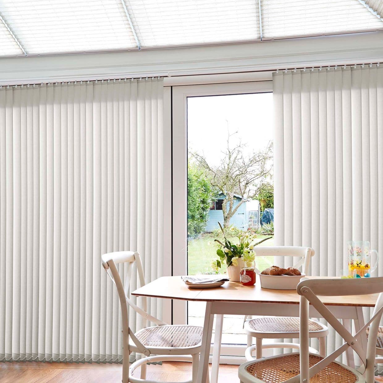 Cream vertical blinds, fully closed across patio doors with a narrow central opening, filtering soft daylight into a bright dining nook with wooden table, chairs, and garden view.