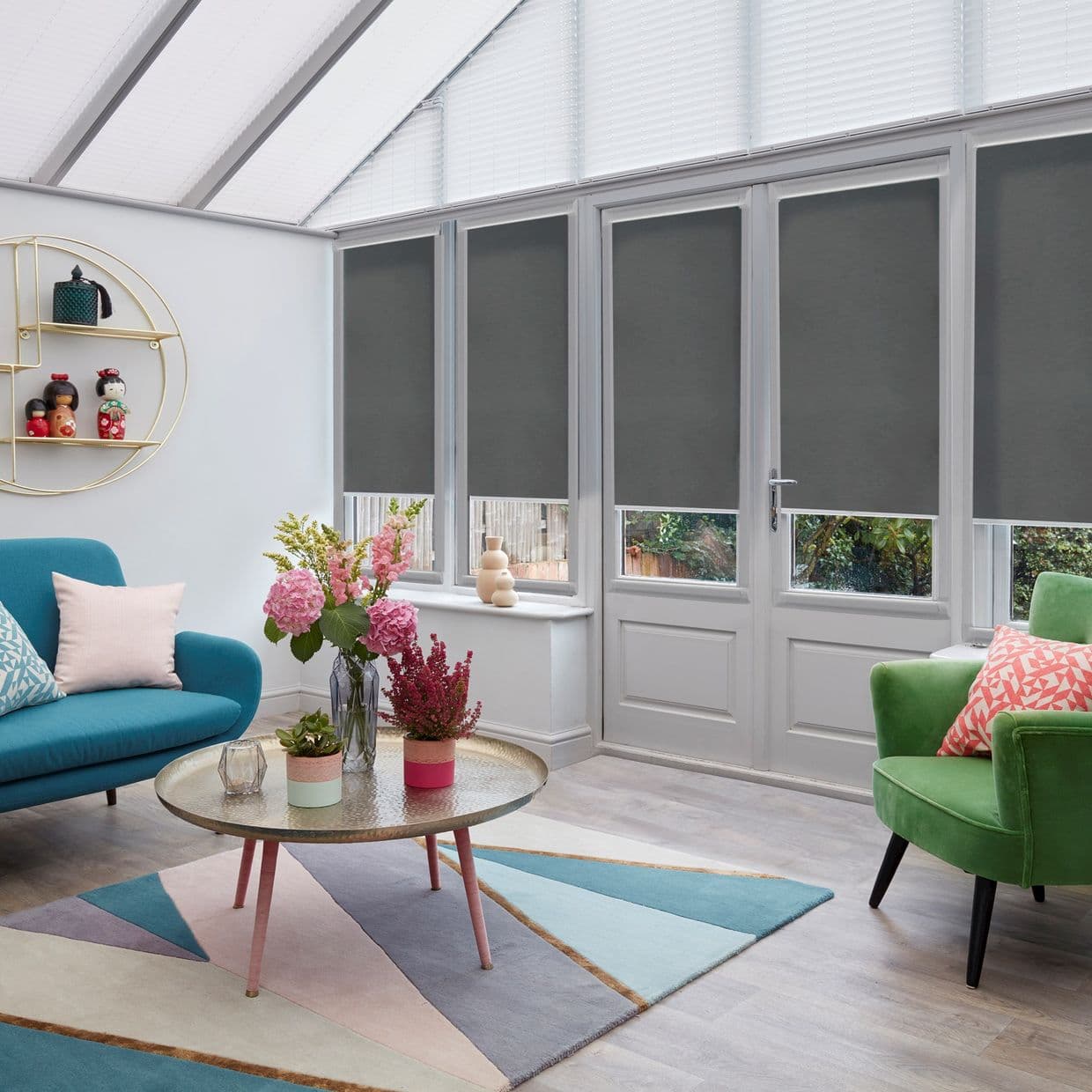 Dark gray roller blinds on tall windows and door, lowered mid-height with pleated blinds on the sloped roof, blocking light; conservatory living area with colorful sofas, rug, and coffee table.