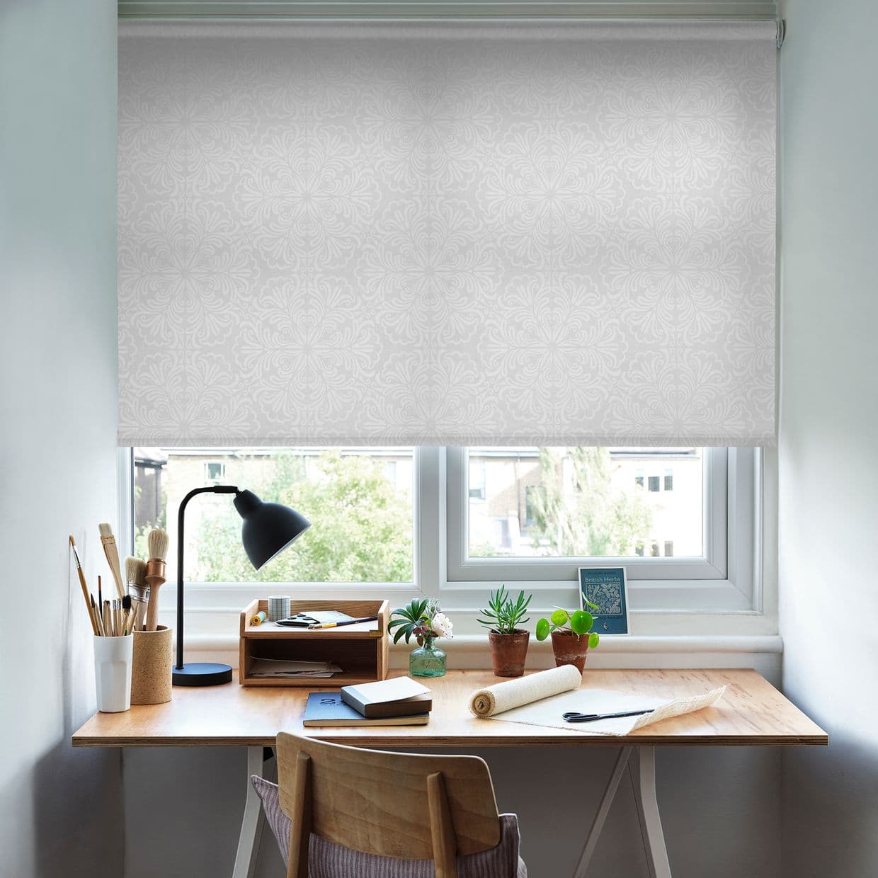 White roller blind with subtle floral pattern, lowered over a double window, filtering daylight; home workspace below has a wooden desk, black lamp, paintbrushes and small potted succulents.