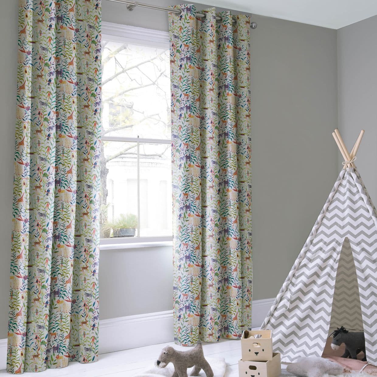 Grommet-top curtains with colorful animal-and-foliage print, hung each side of a sash window on a rod, framing and filtering daylight in a pale-gray children's playroom with a chevron teepee.
