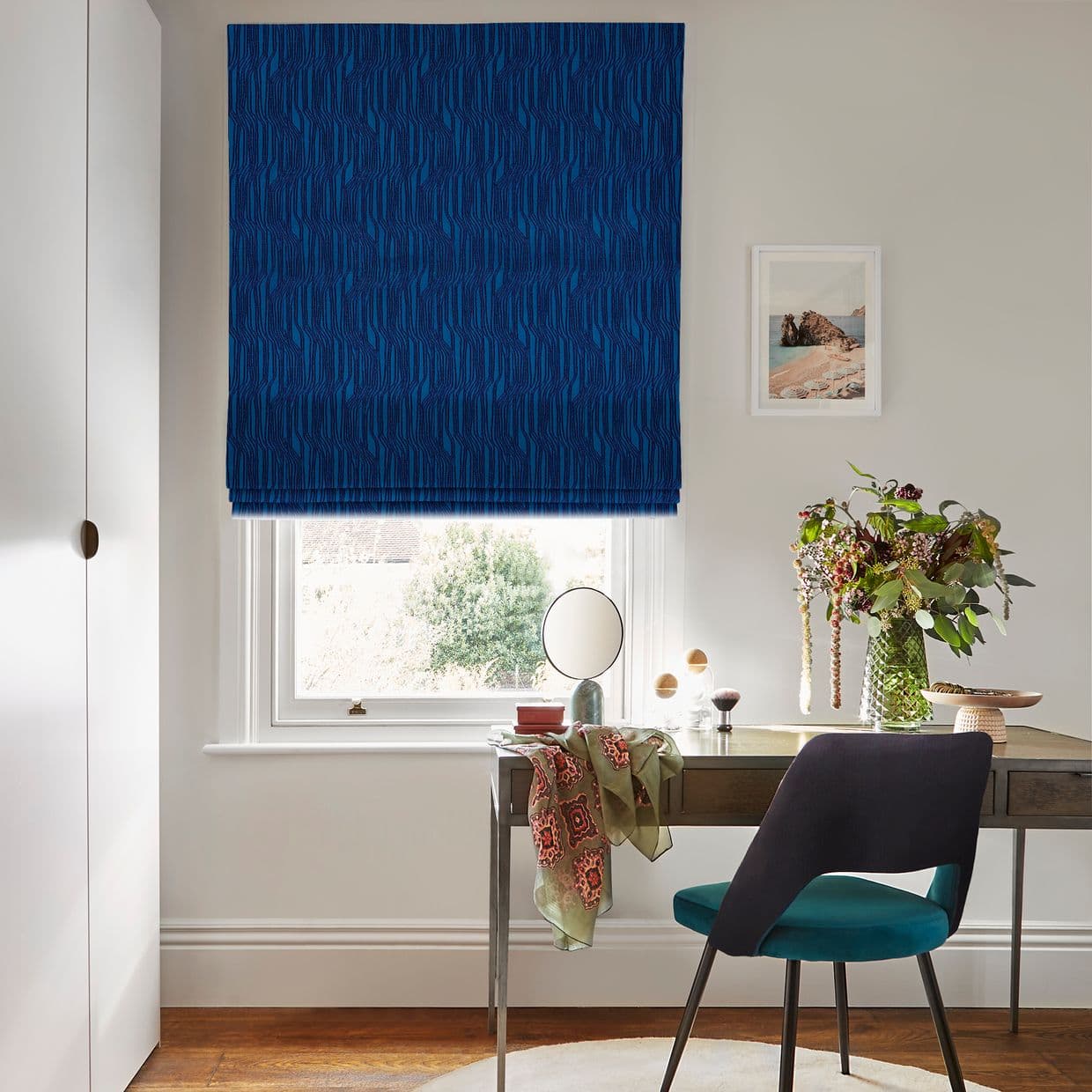 Blue patterned Roman blind, lowered and slightly filtering light; covering a sash window above a desk in a bright home office with teal chair, vase of flowers and seascape print.