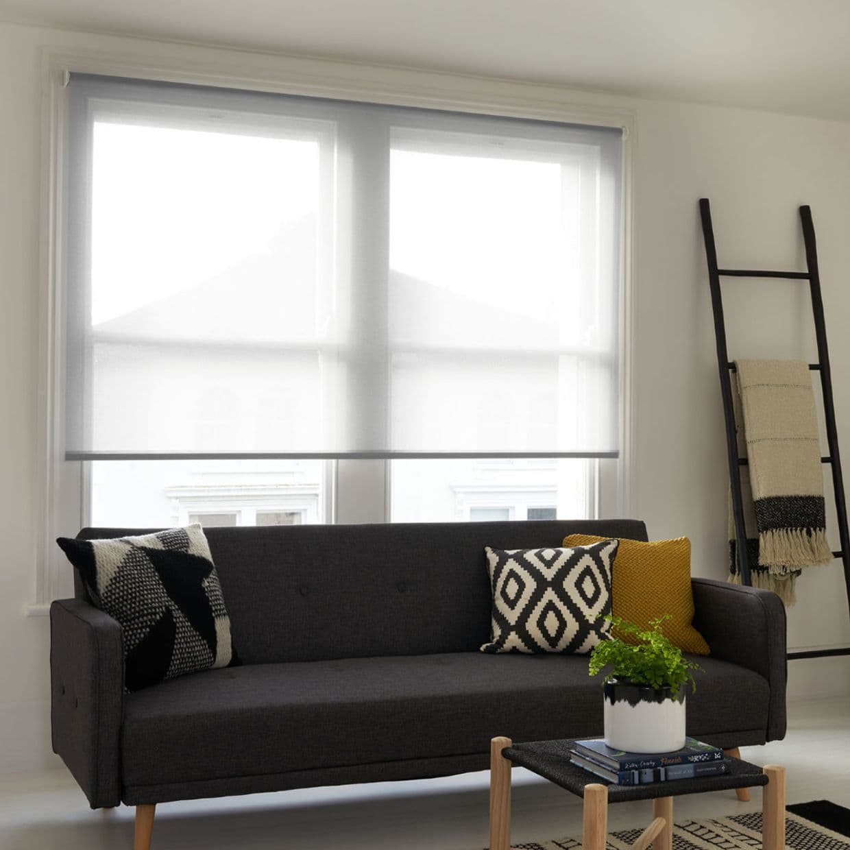 Sheer gray roller blind, fully lowered across a double window, gently diffusing daylight; modern living room with dark gray sofa, patterned cushions, leaning blanket ladder, small coffee table and plant.