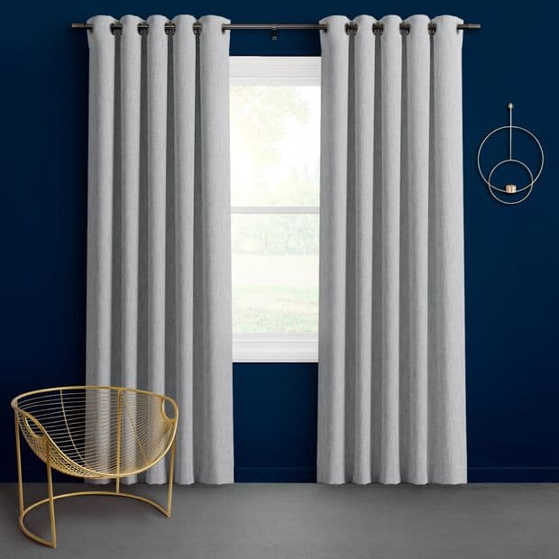 Light-gray grommet-top curtains frame a central window, parted to reveal bright outdoor light; set against deep navy walls with gray floor and a gold wire accent chair to the left.
