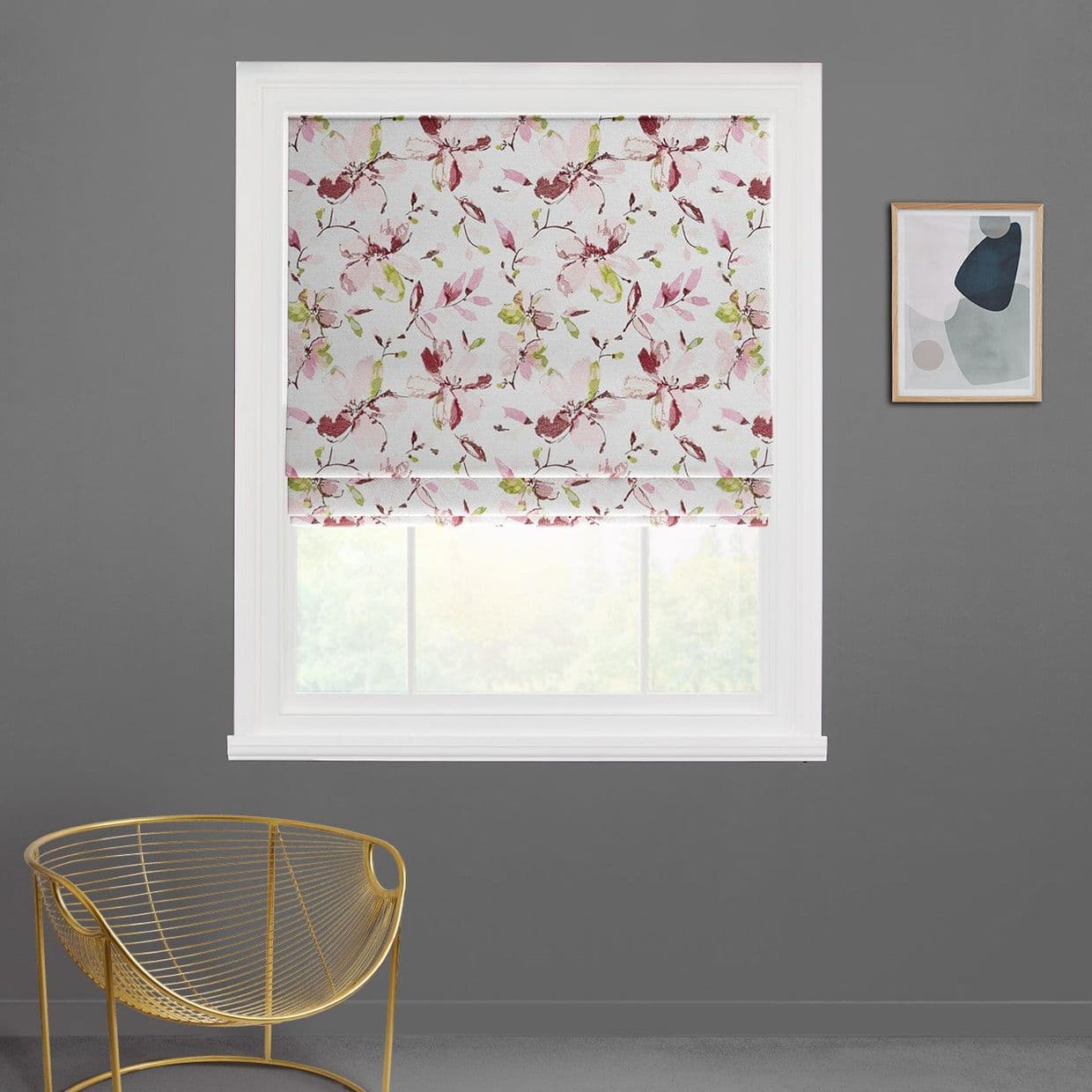A white Roman blind with pink-red floral and green leaves, partially lowered and softly folded, filtering daylight through a white-framed window; grey wall, gold chair, abstract print.