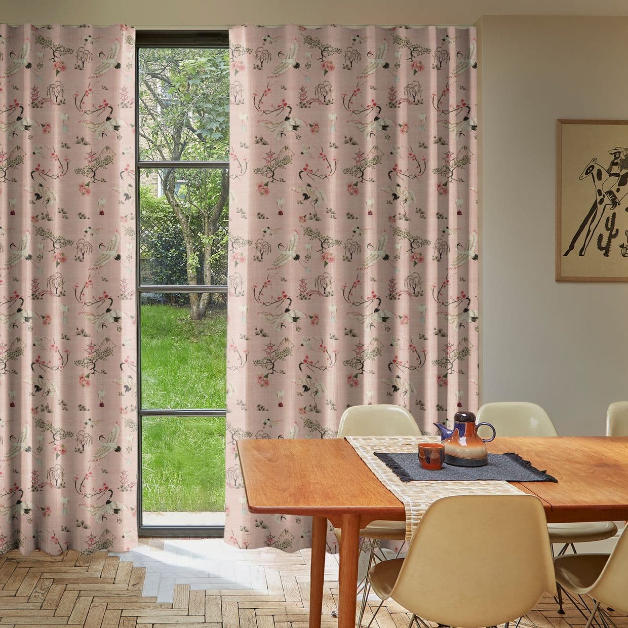 Floor-length pink patterned curtains (crane and blossom motif) hung either side of a tall black-framed window, drawn mostly closed with a central gap, filtering soft daylight into a dining room.