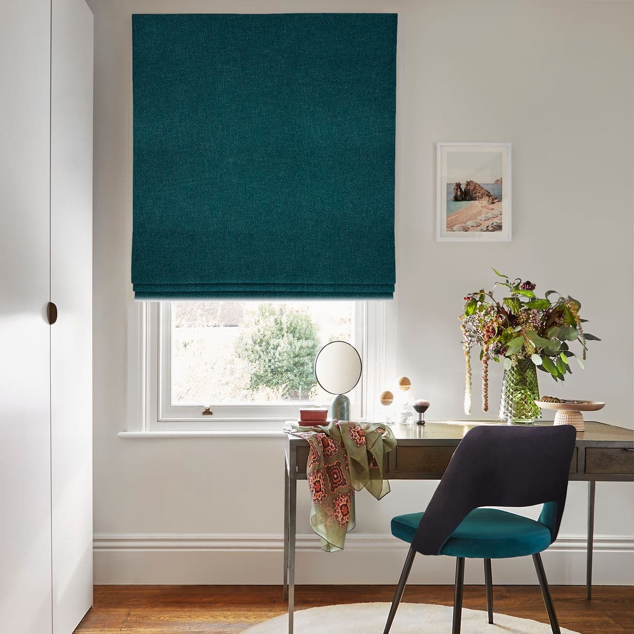 Teal Roman blind, lowered to cover most of the window with folded hem; filtering soft daylight; above desk with chair, vase of flowers, mirror, pale walls and garden view.