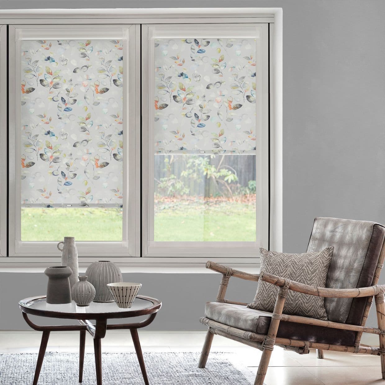 Two translucent roller blinds with watercolor leaf print cover a double window â€” left down, right partially raised, filtering soft daylight in a neutral living room with rattan armchair, round table.